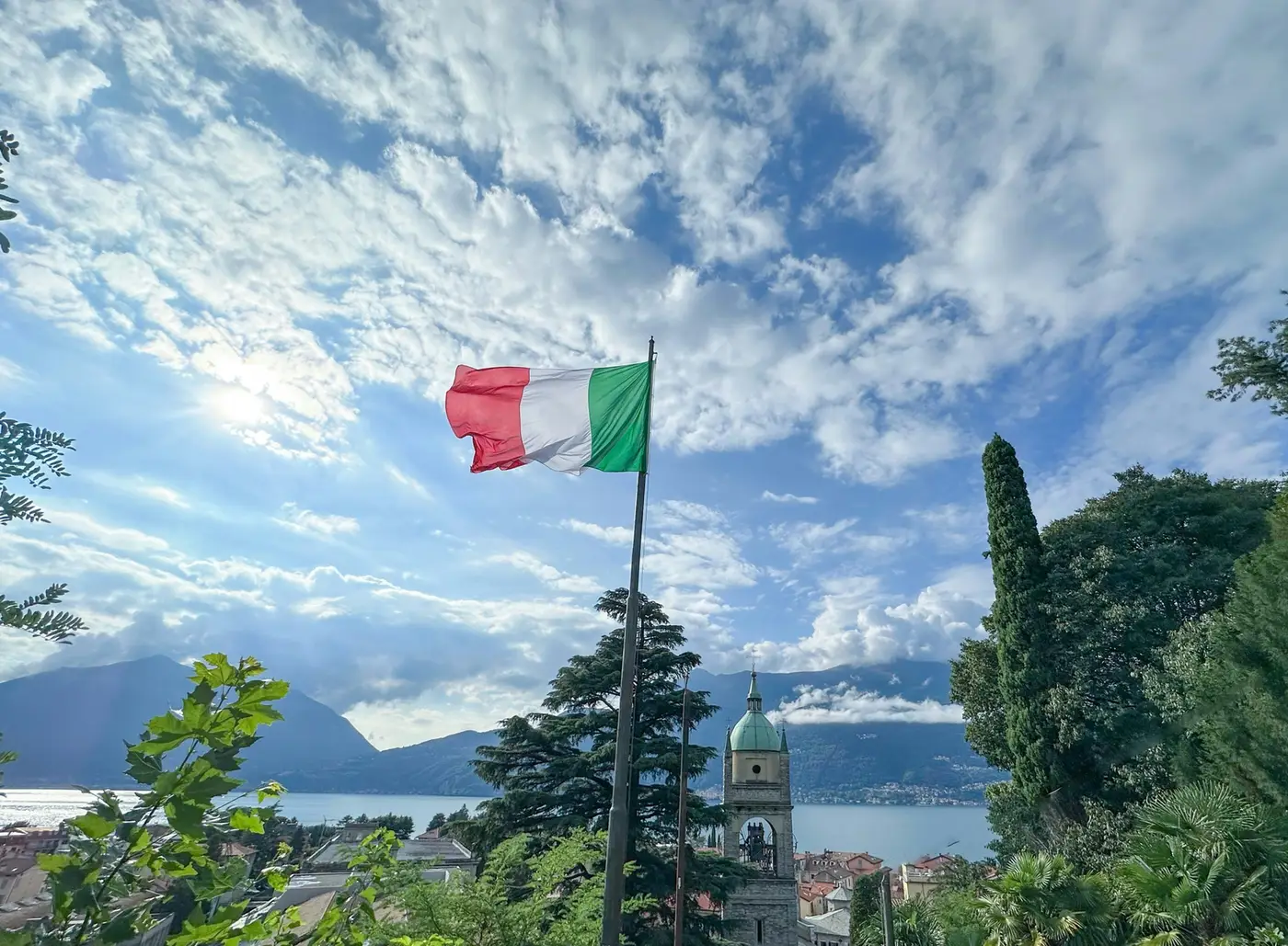 The Italian flag flying over a lakeside town, church tower and mountains beyond