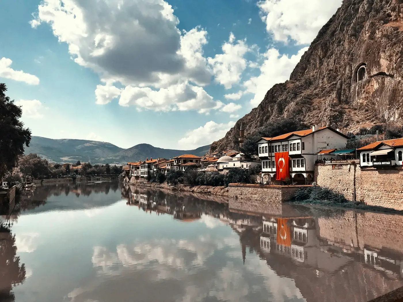 Ottoman-era houses along a river beneath rock cliffs in Anatolia, a Turkish flag flying from a balcony